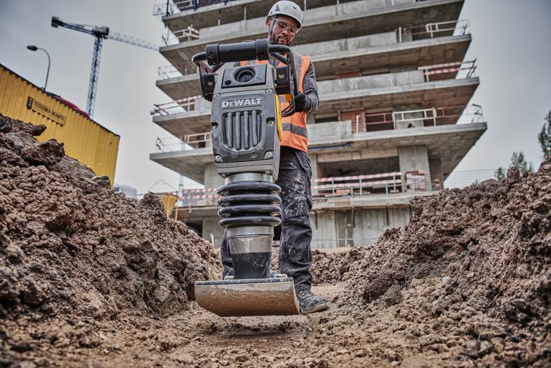 DEWALT compactor gebruikt op bouwplaats, bediend door medewerker in veiligheidsuitrusting, omringd door aarde en gebouwen.