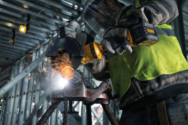 Close-up van een persoon met beschermende handschoenen en veiligheidshesje, die met een draadloze DEWALT-haakse slijper metaal snijdt, terwijl er vonken vliegen op een industriële bouwplaats.